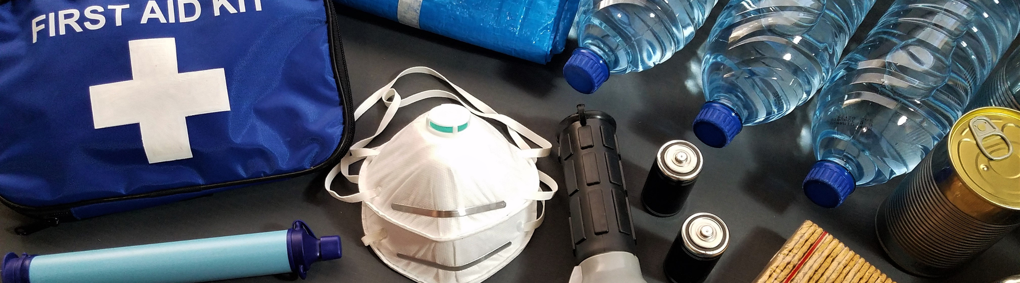 Emergency preparedness kit items arranged on a dark surface, including a blue first aid kit bag with a white cross, several clear plastic water bottles with blue caps, a white face mask, a tourniquet, batteries, and other survival gear.