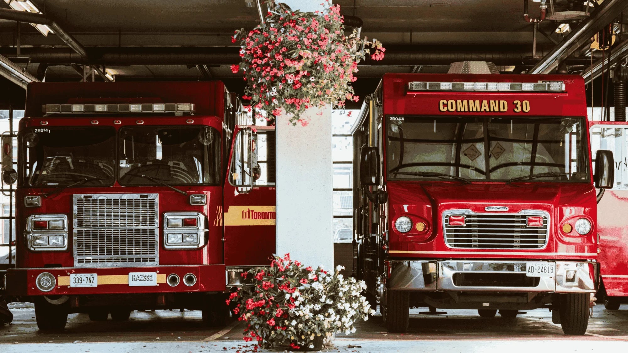 Two red fire trucks parked side by side inside a fire station garage, with hanging flower baskets and floral arrangements in the foreground.